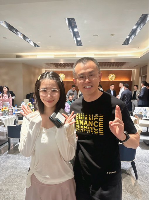 Two people pose for a photo at a YZI incubator meetup—the woman in a white top flashes a peace sign while holding a card reader, and the man beside her, identified as CZ, wears a black "BINANCE" t-shirt and smiles while making a peace sign, with a banquet hall full of attendees visible behind them. The caption playfully references meeting CZ in person and celebrates the incubator's community gatherings.