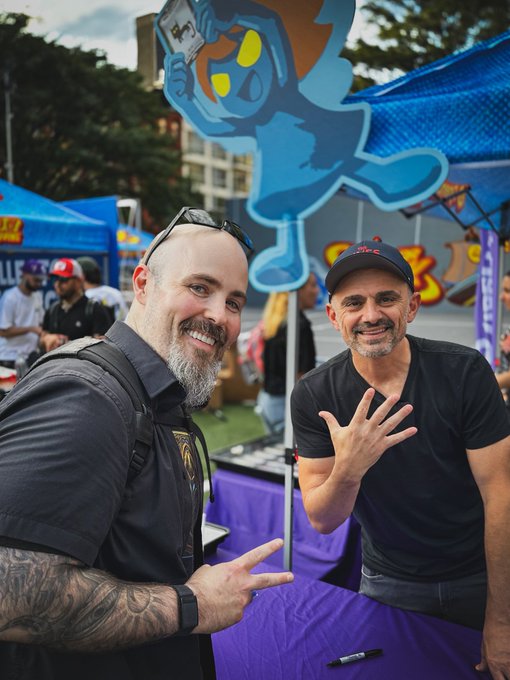 Two men smile at the camera and hold up their hands at an outdoor event, standing in front of a blue tent and a large inflatable blue mascot holding a card.