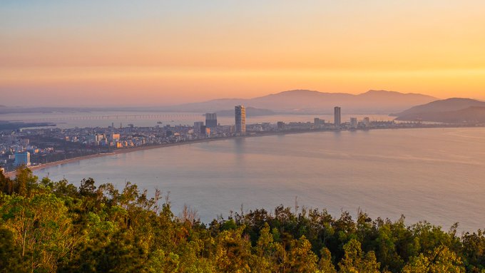 Scenic sunset view of a coastal city with mountains in the background, taken from an elevated vantage point overlooking the bay and shoreline.