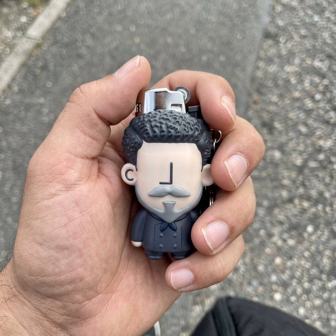 A hand holds a small figurine of a stylized character with gray curly hair, a mustache, and a dark coat, functioning as a lighter.