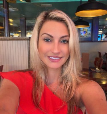 A blonde woman with blue eyes smiles brightly at the camera in a selfie taken inside what appears to be a restaurant or cafe, wearing a vibrant red one-shoulder top with ruffled details.
