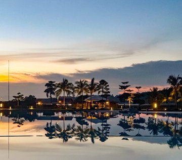 A serene sunset/sunrise scene over a calm body of water, with palm trees silhouetted against a colorful sky reflecting perfectly on the water's surface.