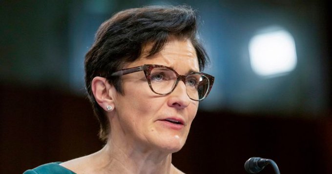 A close-up photograph of a woman with short brown hair and tortoiseshell glasses speaking into a microphone, appearing to be in a formal hearing setting.