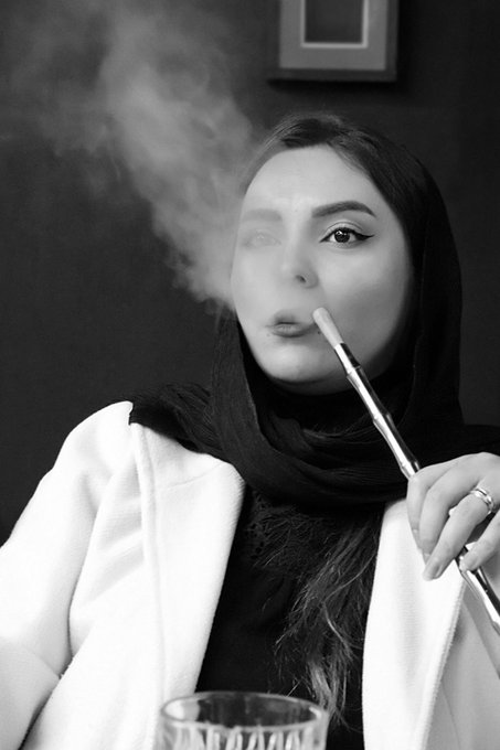 A black-and-white photograph shows a woman in a dark headscarf and white coat exhaling smoke from a long hookah pipe, her gaze directed at the camera, embodying the contemplative, melancholic mood suggested by the title "Hidden Sorrows" from the XTZ collection on Objkt.