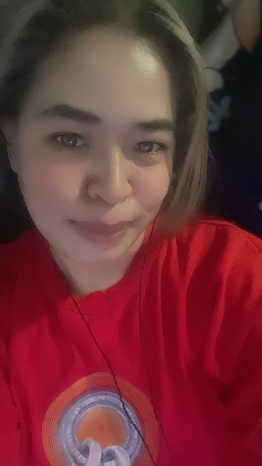 Selfie of a young woman with brown hair wearing a red shirt and earphones in a dimly lit setting.