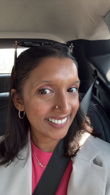 A smiling woman with dark hair wearing a nose ring, hoop earrings, and a pink top under a gray blazer, seated in a car with a seatbelt on, with sunglasses perched on her head.