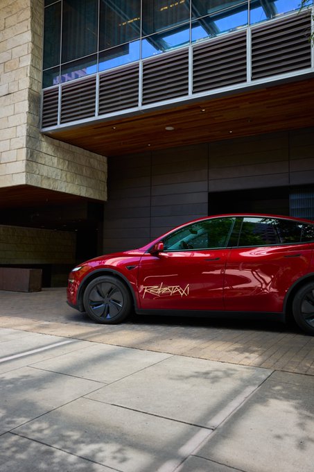 A red Tesla Model Y with "REBAWN" text on its side is parked beneath a modern building's overhang, fitting the "Cybecab launch event" post as a sleek electric vehicle suitable for an innovative transportation debut.