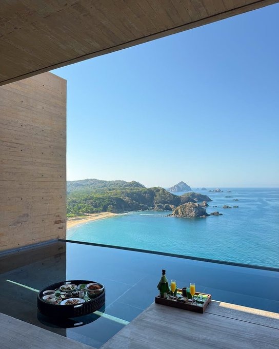 A scenic view from a modern, concrete-walled room looking out over an infinity pool toward a beach, rocky coves, and the ocean under a clear blue sky, with a breakfast tray and a bottle of champagne on the ledge.