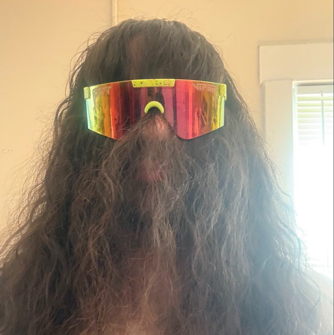 A bearded person with long, flowing brown hair that completely covers their face wears reflective rainbow-tinted visor sunglasses, humorously using their hair to literally block their view — a playful visual gag matching their caption about avoiding the urge to check the charts.