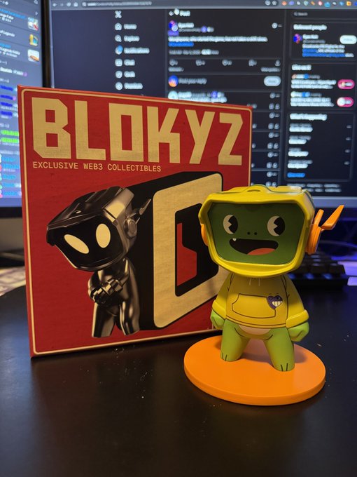 A green Blokyz figurine in a yellow hoodie stands next to its red "BLOKYZ - Exclusive Web3 Collectibles" box, shown in front of computer monitors. The image supports the post's question about whether figurine holders already have their Blokyz whitelist (WL) spot.