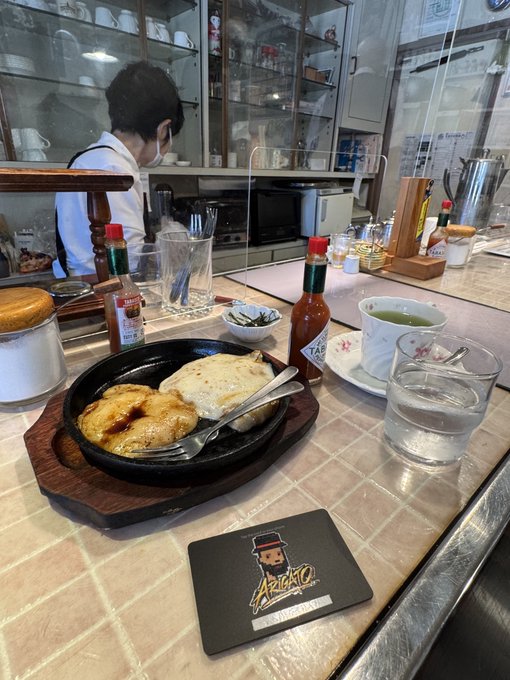 A photo taken at a diner counter shows a sizzling plate of food with eggs and fried items, a cup of green tea, hot sauce, and a coaster with a character logo labeled 'ARIGATO.'