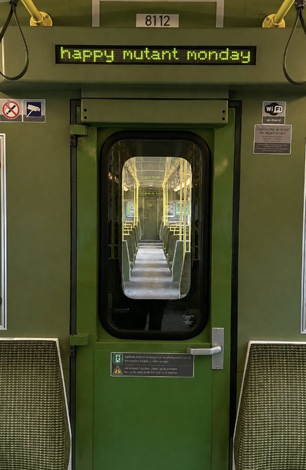The interior of a green transit train car (number 8112) displays a scrolling LED sign reading "happy mutant monday" above the doorway, fitting the "HMM" abbreviation for the recurring meme greeting.
