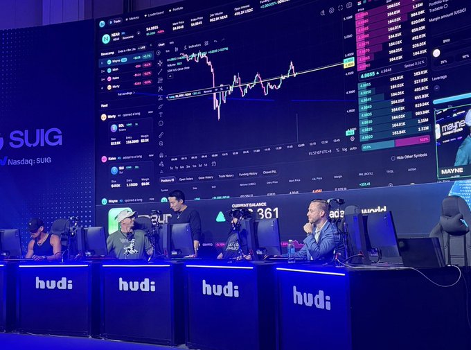 An esports tournament stage with players seated at desks branded 'hudi', in front of a massive screen displaying cryptocurrency trading charts and the ticker SUIG.