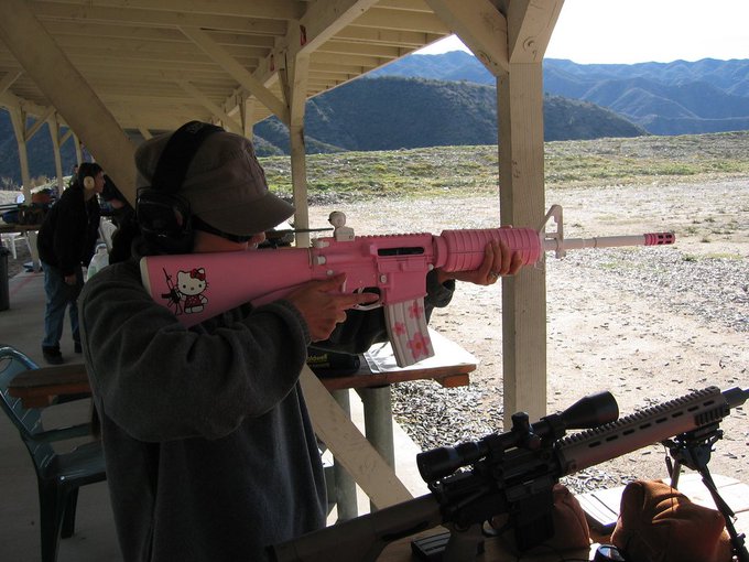A young person at an outdoor shooting range aiming a pink Hello Kitty-themed rifle downrange at the mountainous desert terrain.
