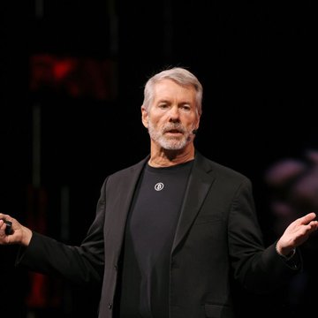The image features a man wearing a black suit, standing on stage with his arms outstretched as if he's giving a speech or presentation. The caption below the image reads "LATEST- @MicroStrategy Sold 32 #XRP". This suggests that the man might be discussing a recent event involving cryptocurrency, possibly related to MicroStrategy and Bitcoin.