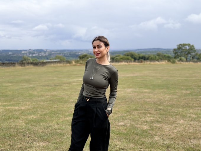 A young woman with brown hair stands in a field wearing an olive green long-sleeve top, black pants, and a heart-shaped necklace.