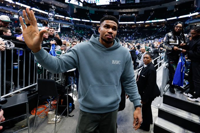 NBA player Giannis Antetokounmpo wearing a blue 'BAYC' hoodie waving at fans while walking through a basketball arena tunnel.