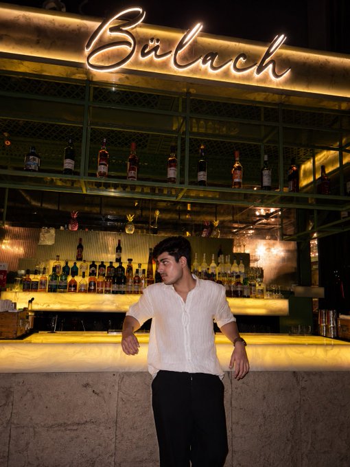 A young man in a white shirt and black pants stands at the bar counter of 'Baladh', leaning against the stone ledge beneath an illuminated sign and shelves lined with liquor bottles.