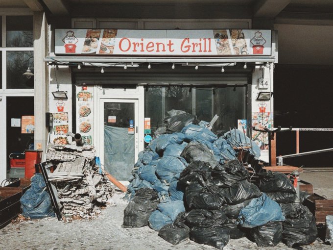 The storefront of 'Orient Grill' is shown piled high with black and blue garbage bags, snow, and debris outside its door.