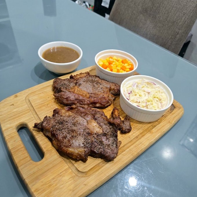 A photograph of a meal on a wooden cutting board featuring two grilled pork steaks, a bowl of brown gravy, a bowl of corn, and a bowl of coleslaw on a glass table.