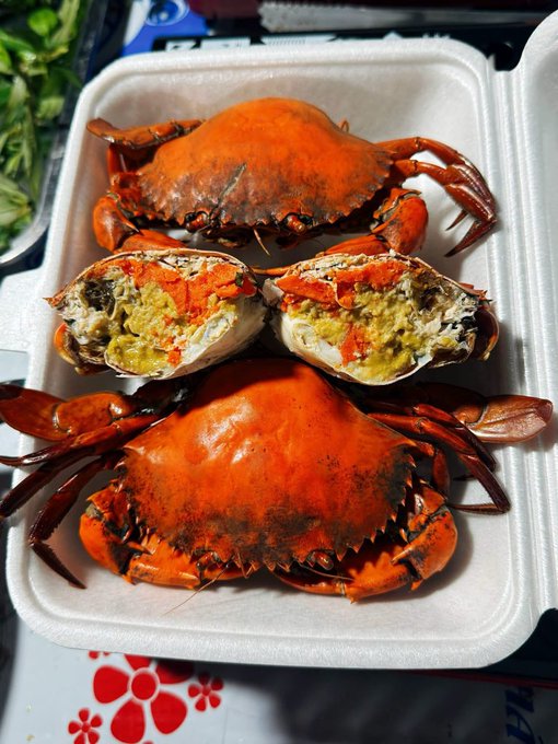 A takeout container filled with cooked orange crabs, including one split open revealing yellow and orange roe and meat inside.