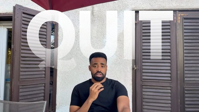 A man with a beard in a black t-shirt sits outdoors in front of brown louvered shutters with the word "QUIT" overlaid in large translucent white letters across the top of the frame.