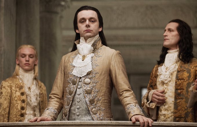 A pale, aristocratic vampire with long dark hair stands solemnly in an ornate gold-embroidered jacket, flanked by two similarly dressed figures in elaborate baroque attire, his expressionless gaze and rigid posture evoking the formal "approval" stance referenced in the caption. The scene appears to be from the film *Interview with the Vampire*, capturing Louis in a moment of regal judgment fitting the post's theme of final approval.
