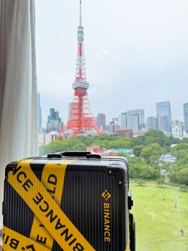 A black Binance-branded suitcase with yellow Binance tape sits in the foreground, with the Tokyo Tower and city skyline visible through a window in the background.