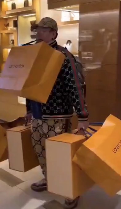 A man in a Gucci-patterned tracksuit and cap is loaded down with multiple large orange Louis Vuitton shopping bags and boxes inside what appears to be a luxury boutique. The caption humorously suggests this scene will become a memorable, legendary moment referenced in the future.