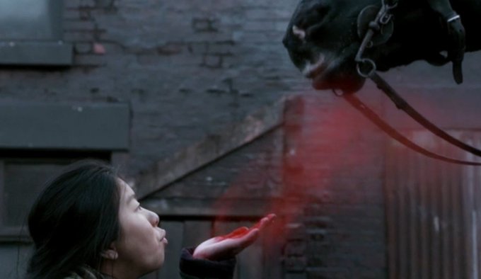A scene showing a young woman extending her hand toward a snarling, chained creature as red mist floats between them, set against a dark brick wall.