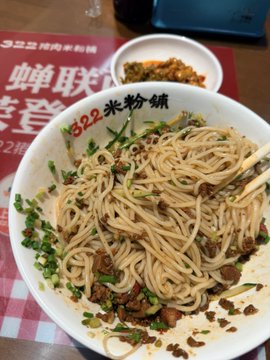 The image shows a bowl of noodles with meat and vegetables sitting on top of a table. The noodles appear to be overcooked or burnt, indicating that they have been left unattended for quite some time. The bowl has a red background, which contrasts with the white color of the noodles. In the background, there is a plate containing more food items, suggesting that this meal was meant to be shared among multiple people.