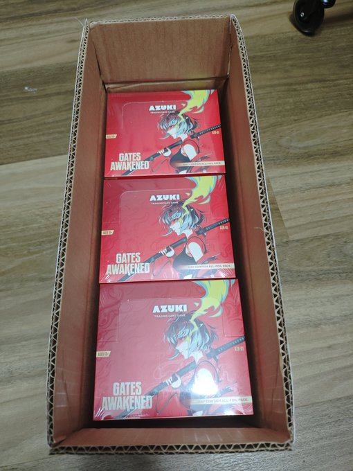 Three red 'Gates Awakened' Azuki Trading Card Game booster boxes stacked inside a cardboard shipping box on a wooden floor.