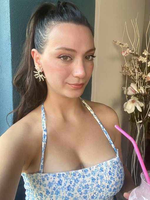 A young woman with dark hair in a ponytail poses for a selfie, wearing a blue floral halter top and starburst earrings, with a pink straw drink and flowers visible.