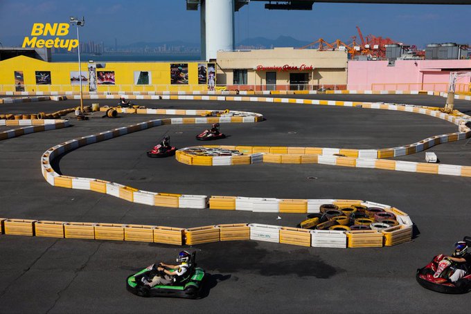 An outdoor go-kart track with yellow and white barriers where several people are racing karts, with 'BNB Meetup' watermarked in yellow text on the upper left.