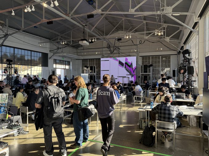 A large industrial warehouse space filled with people at tables working on laptops, with a person in the center wearing a 'Team' shirt, and stage lighting set up along the ceiling.