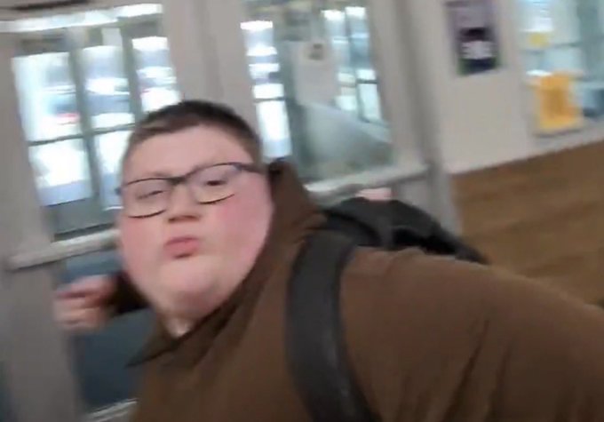A blurry selfie-style photo of a young person with glasses wearing a brown hoodie and backpack in what appears to be a hallway or public building.