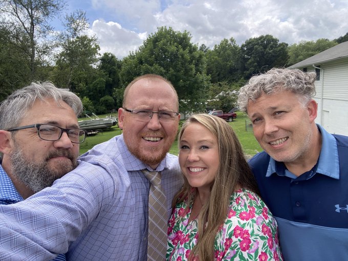 A selfie of four people—three men and one woman—smiling outdoors with trees and a house in the background.