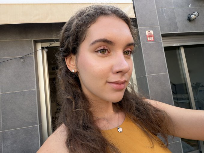 A close-up selfie shows a young woman with long wavy brown hair, green eyes, and a yellow top, standing in front of a tiled wall and glass doors.