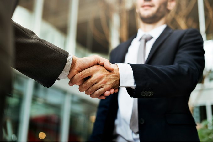 Two businessmen in dark suits shake hands in front of a glass-fronted building, symbolizing the formal agreement between 39 state banking associations forming the BankChain Alliance to develop bank-owned blockchain infrastructure for tokenized deposits, stablecoins, and automated settlement, with a targeted 2027 launch.