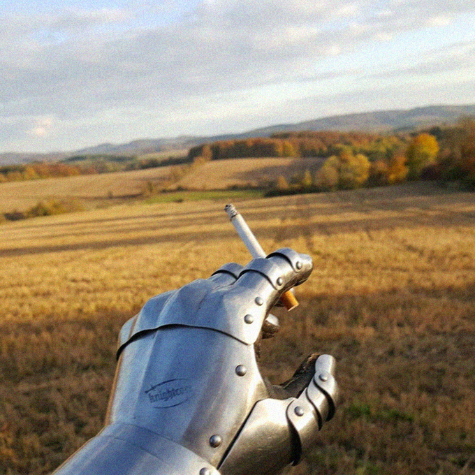 A metal gauntlet with riveted plates points toward a scenic landscape of rolling hills, autumn-colored trees, and a harvested field under a cloudy sky.