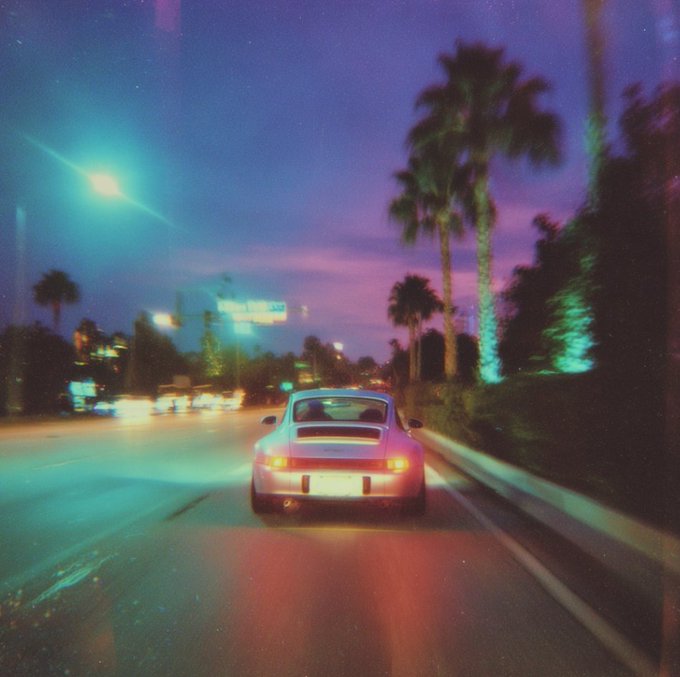 The image shows a car driving down a street at night, with palm trees lining both sides of the road. The car appears to be a sports car, possibly a Porsche, as indicated by its design. The photo has been edited or filtered, giving it a purple tint and making the sky appear pinkish-purple in color. This gives the scene an artistic and dreamy feel, with the car driving away from the viewer on the right side of the image.
