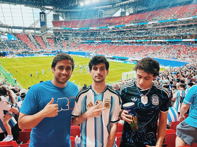 Three young men pose together in the stands of a large soccer stadium; one wears an Argentina jersey, another wears an Argentina jersey, and the third holds a cup while wearing an Argentina jersey.