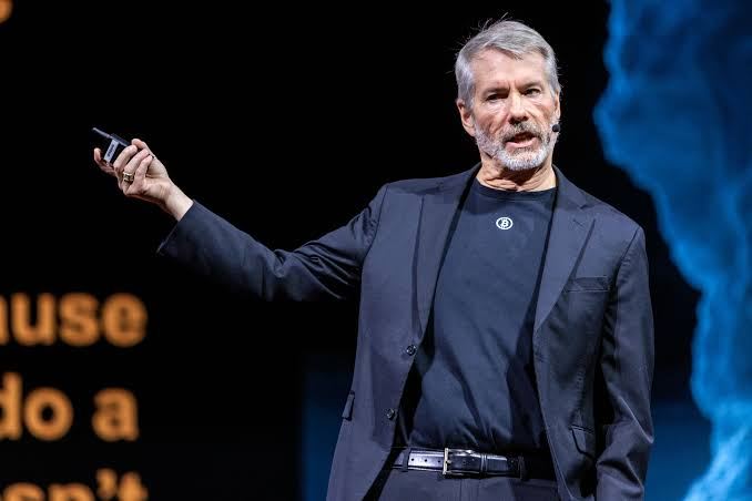 A man with a gray beard wearing a dark suit and t-shirt speaks on stage while holding up a clicker device, gesturing with one arm raised. Partial text visible in the background reads "use," "do a," and "sn't," suggesting presentation slides. The image appears unrelated to the post's content about Strategy-linked Bitcoin wallet transfers, though it may depict Michael Saylor delivering a presentation on cryptocurrency.