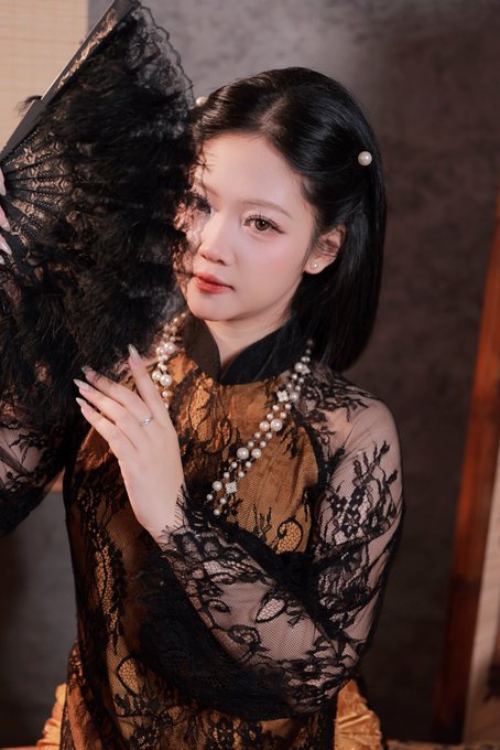 A young woman with long black hair styled with pearl accessories poses elegantly, holding an ornate black lace fan that partially veils her face. She wears a richly textured amber and black lace dress with a pearl-trimmed collar, paired with a layered pearl necklace and delicate rings, while soft warm lighting highlights her serene expression. The vintage-inspired portrait complements the cheerful "Good morning" post, offering a graceful and stylish start to the day.