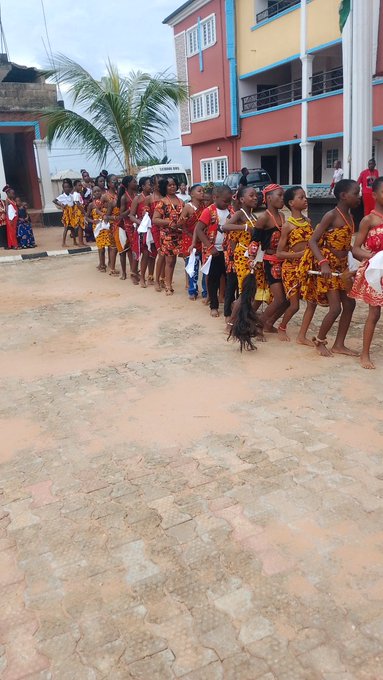 A group of young women in matching traditional African attire, featuring red and yellow patterns, walk in a line on a paved area near buildings.