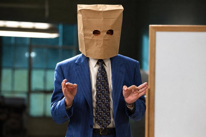 A man in a blue pinstripe suit and patterned tie stands in what appears to be an office or presentation room, wearing a brown paper bag over his head with two eye holes cut out, gesturing with his hands as if explaining something next to a whiteboard—visually embodying the post's sentiment of blind faith in a token with questionable "ponzinomics," where investors buy in without questioning the fundamentals, much like purchasing $wif early.