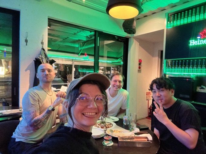 A group selfie of four people smiling at a restaurant table with food and drinks, with a green-lit Heineken sign in the background.