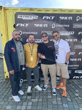 The image shows a group of four men posing for a photo, standing next to each other against a yellow wall with various logos such as "FTK", "Other Page", and "Take me back to ApeFest". The men are wearing different colored clothing - one is in a blue jacket, another has a yellow shirt, the third is wearing a black t-shirt, and the fourth is dressed in white. They all have name tags on their shirts, indicating that they might be attending an event or gathering related to the logos displayed on the wall. The image captures a moment of camaraderie among these individuals as they pose for a picture together.