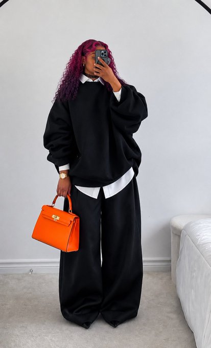 A mirror selfie of a woman with vibrant pink and purple curly hair wearing an oversized black sweatshirt and wide-leg black pants over a white collared shirt, holding an orange Hermès Birkin-style handbag.