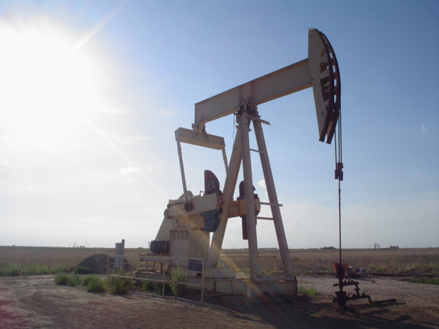 An oil pump jack silhouetted against a bright sun in a flat, rural landscape.